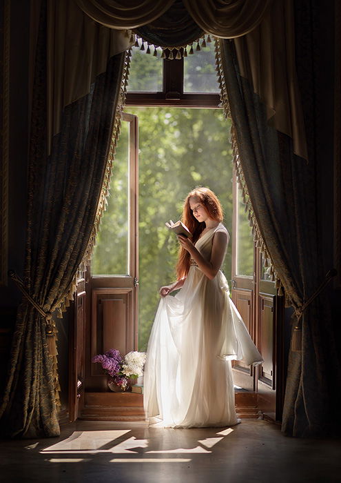 A full-body portrait of a female model reading by curtains and open balcony doors by fine art photographer Olga Fler