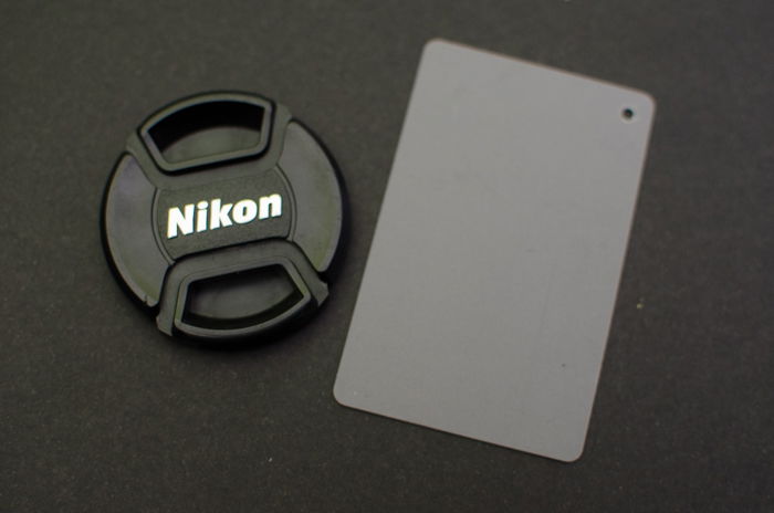 A Nikon lens cap on grey background with a photography grey card beside it