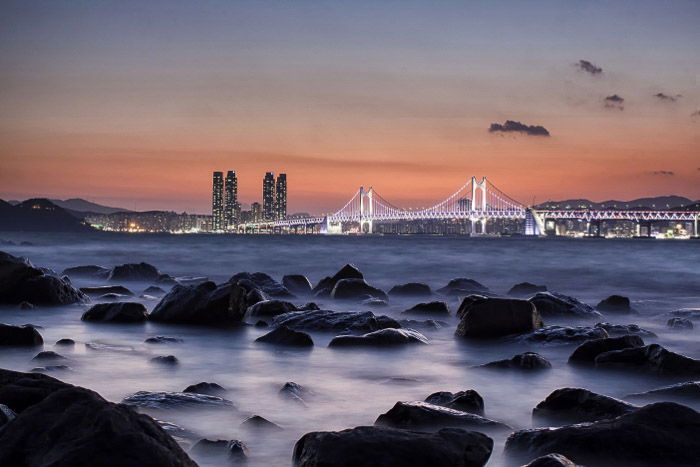 A rocky coastline in the foreground of a coastal cityscape at sunset shot with slow shutter speed