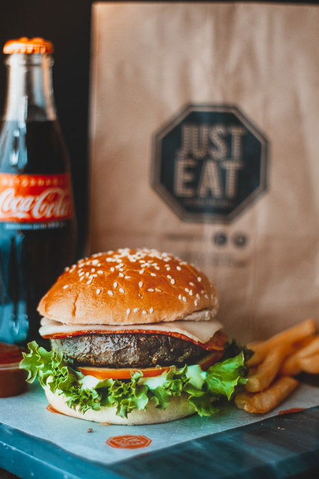 A delicious hamburger beside a softdrink and paper food bag 