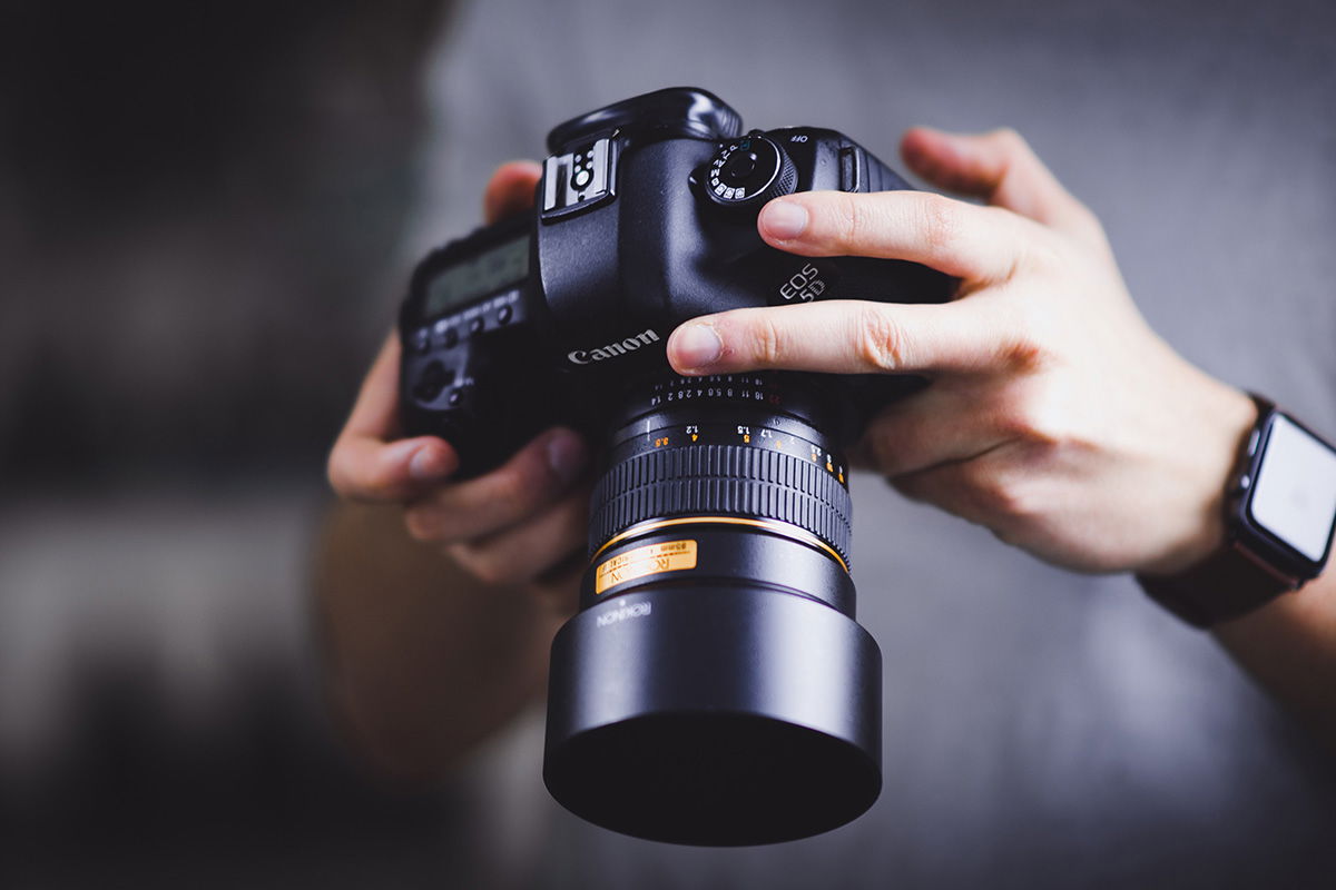 Two hands holding a digital camera and lens downward for a person to look at its back screen