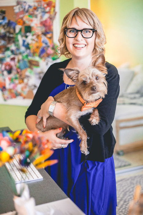 A fun and colorful headshot photo of a woman posing indoors with a small dog