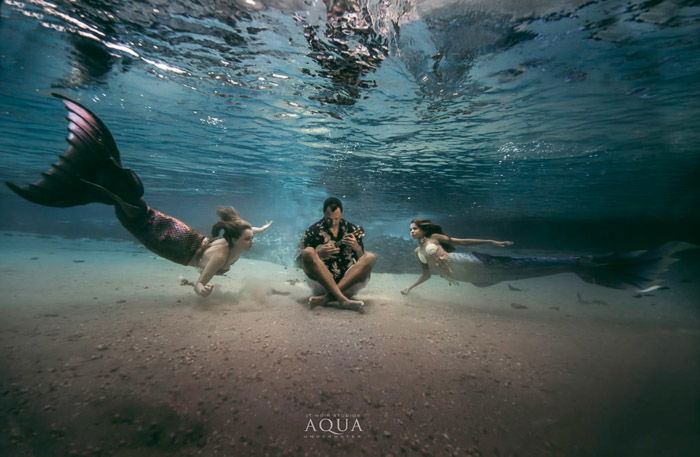 A magical underwater mermaid fantasy scene featuring two mermaids swimming towards an underwater musician - mermaid photoghraphy tips