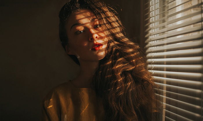 A moody portrait of a female model posed by a window, shadows of the window blinds cast on her face and hair - examples of dark portraits