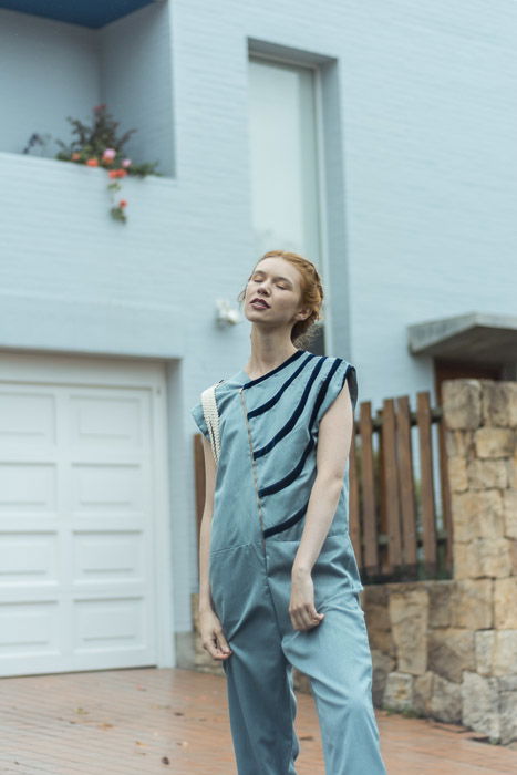 A woman stands in front of a house wearing blue coveralls.