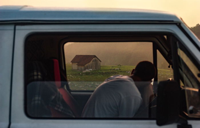 A driver hunched over in a van with a barn in the distance