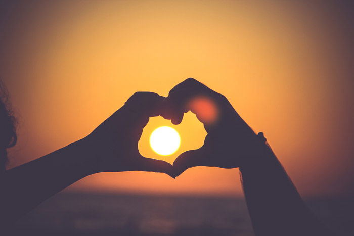 the silhouette of a persons hands making a love heart shape around a setting sun