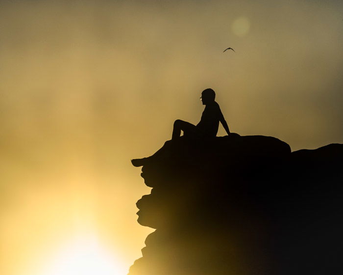 Silhouette of a boy sitting on the edge of a cliff