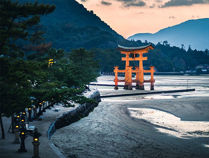 A beach scene at sunset, featuring a wooden torii gate in the foreground and a distant mountain view with mountains lit up by headlights. 