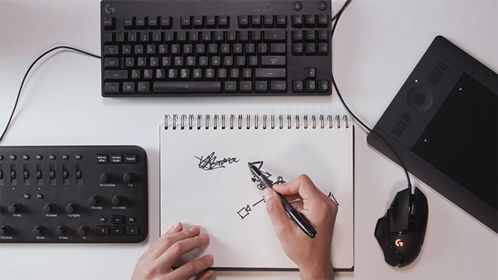 A person writing with a pen on paper next to computer equipment.