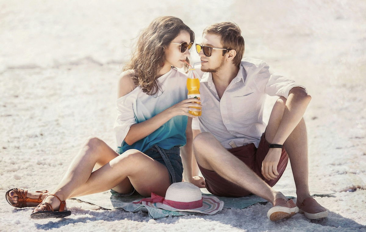 A couple with sunglasses and summer clothes on sitting on a beach their heads turned to each other with a drink held up between them