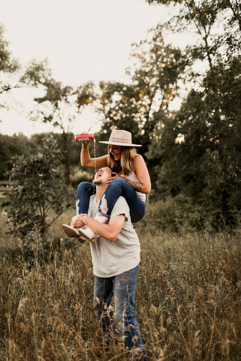 A woman in a hat sitting on a man's shoulders pouring a drink into his mouth for couple poses