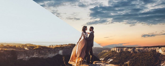 A couple sharing an intimate embrace while standing on a rocky surface at sunset, with the silhouette of mountains and a hilltop in the background. 