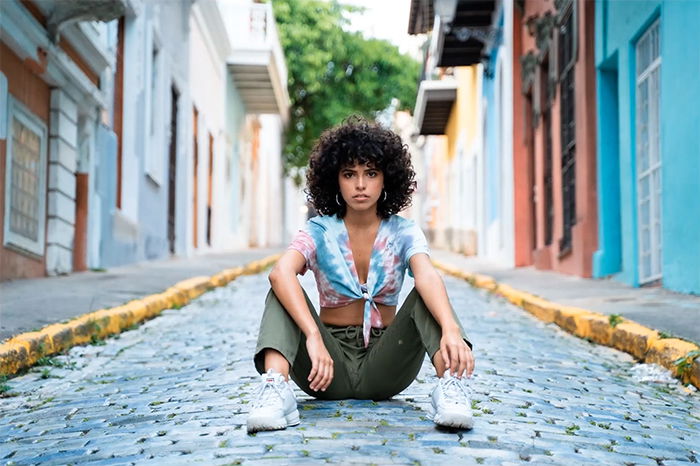 A woman sitting on a stone paved street with buildings in the background, smiling at camera. 