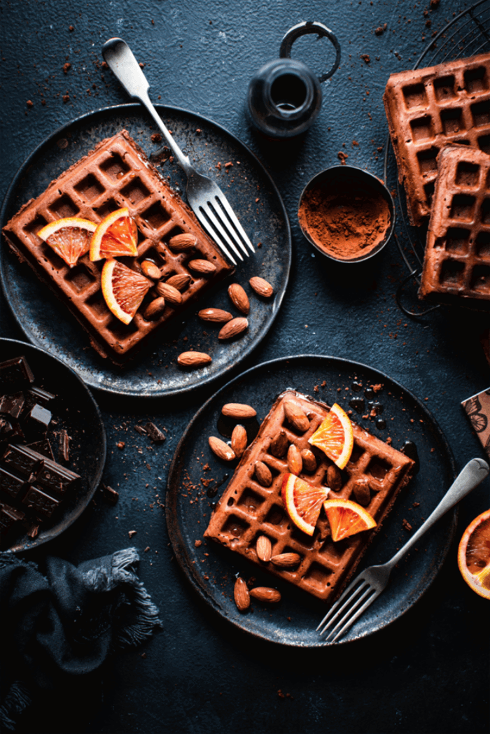 A plate with waffles, topped with orange slices and sprinkled with powdered sugar, surrounded by additional dessert items. 