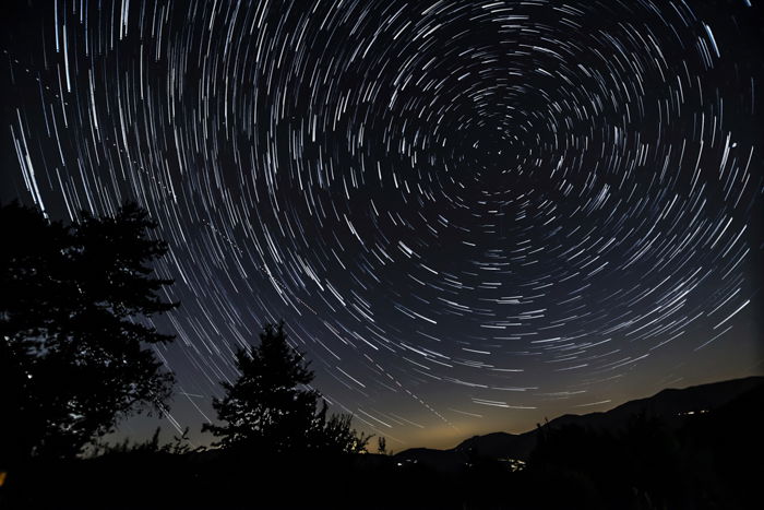 Star trail photograph around the North Polar star with hills, trees and house lights.