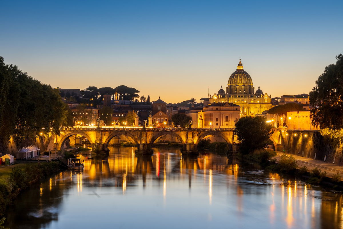 Lit Roman bridge and buildings over a river at dusk with reflections in the water