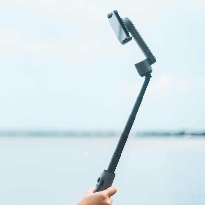 A person taking a selfie with a smartphone and a telescopic camera mount at the beach.