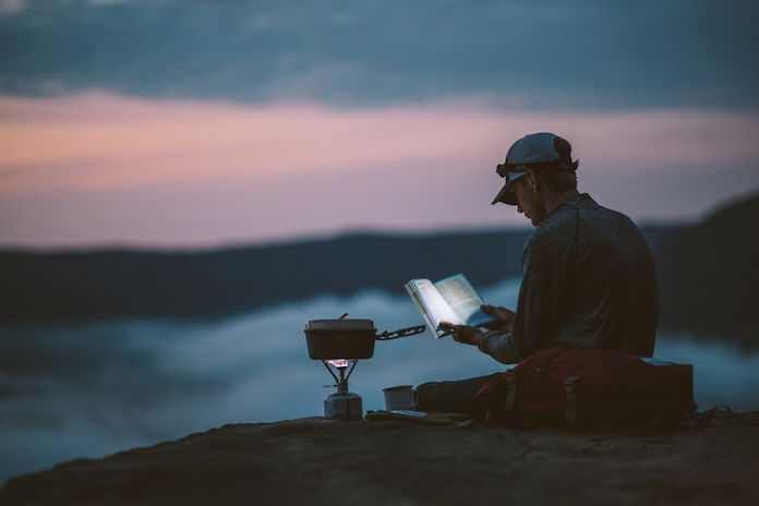 A hiker reading a book outside at dusk on a mountain ridge with a camp stove beside him