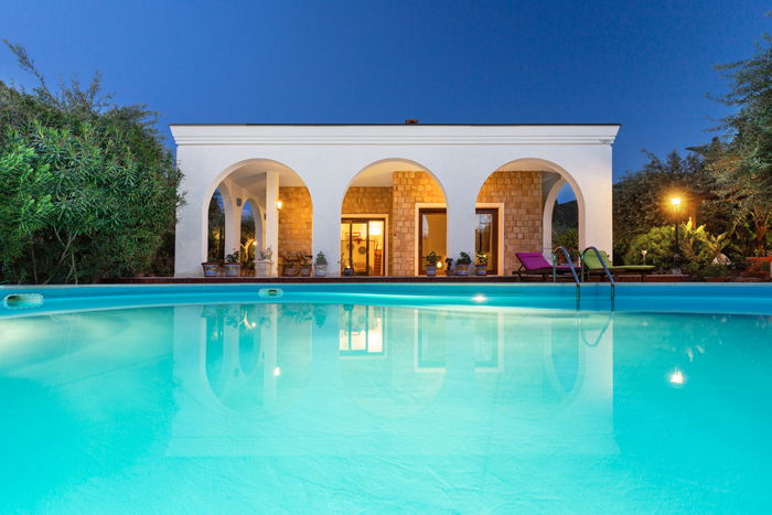 a twilight real estate photography shot of a small pool house with pool in foreground surrounded by bushes