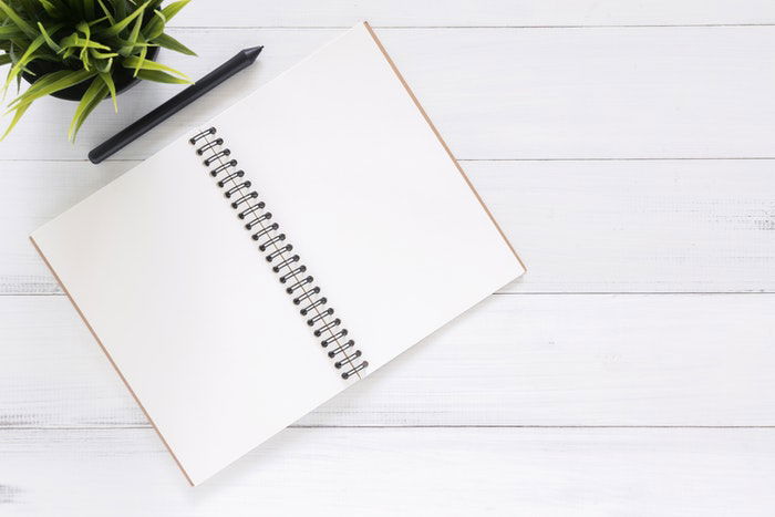 A white, spiral-bound notebook with a black pen on top of it, placed on a wooden surface. 