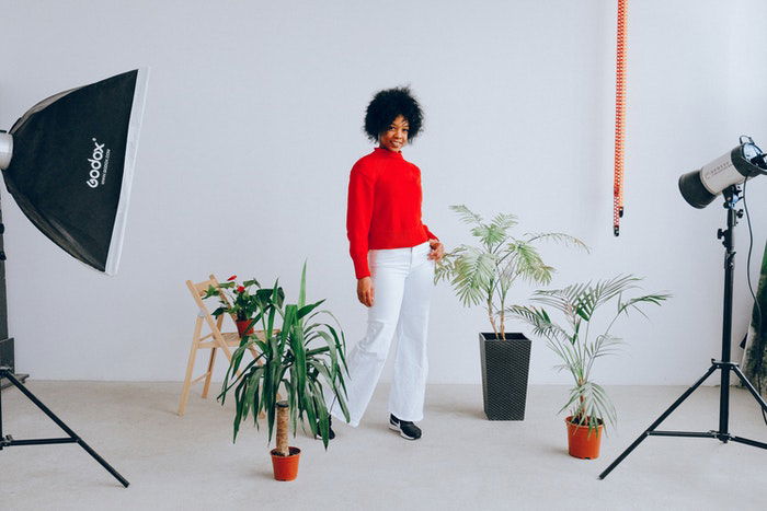 A woman stands in front of potted plants, a white chair, and various photography equipment. 