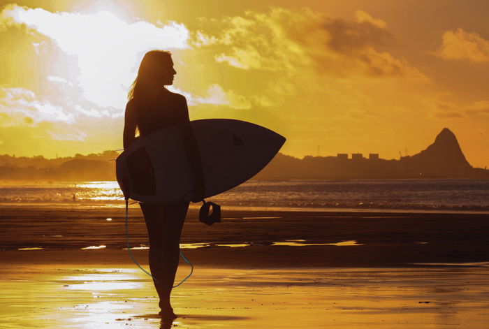 a silhouette of a woman holding a surfboard on the beach with setting sun backlighting