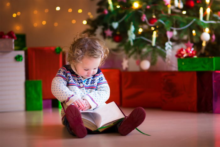 A young child is sitting on the floor in front of a Christmas tree and gifts, holding an open book. 