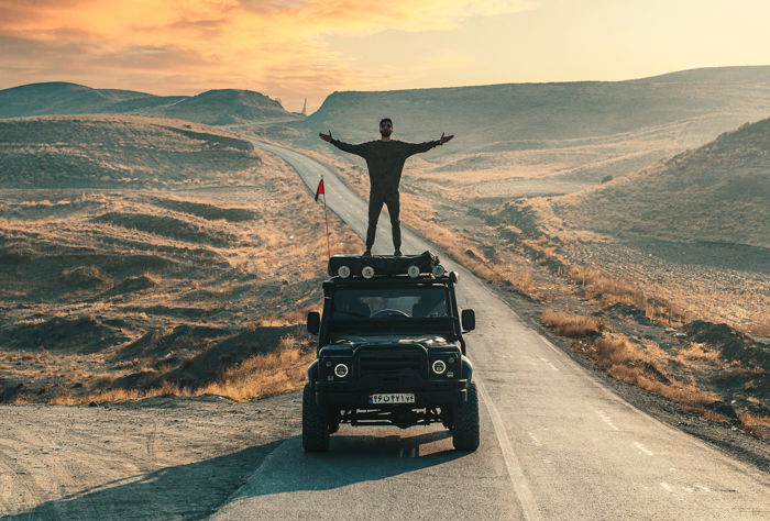 adventure photography of a man standing on top of a truck while driving through the desert