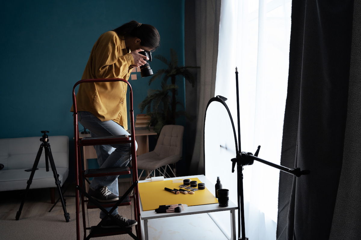 Photographer taking DIY product photography overhead shot from a ladder in a home studio using natural window light