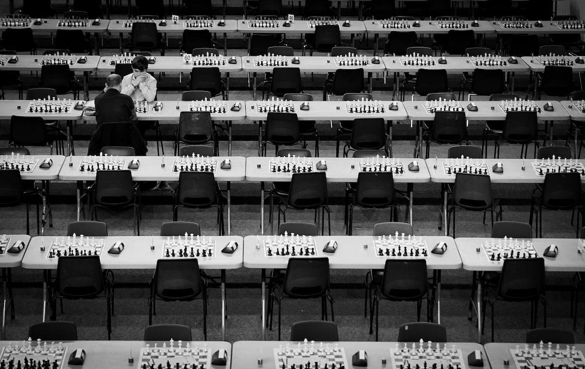 Two chess players among rows of tables, chairs, and chess boards in a repetitive pattern for visual weight