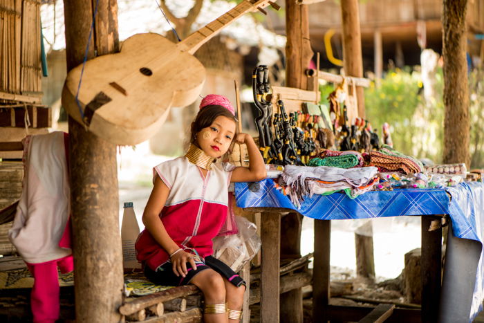 An environmental portrait photography shot of a young Kayan girl beside the table of trinkets she sells outside the family home