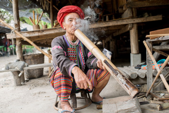 An environmental portrait of a Lahu man outside his house smoking long bamboo pipe