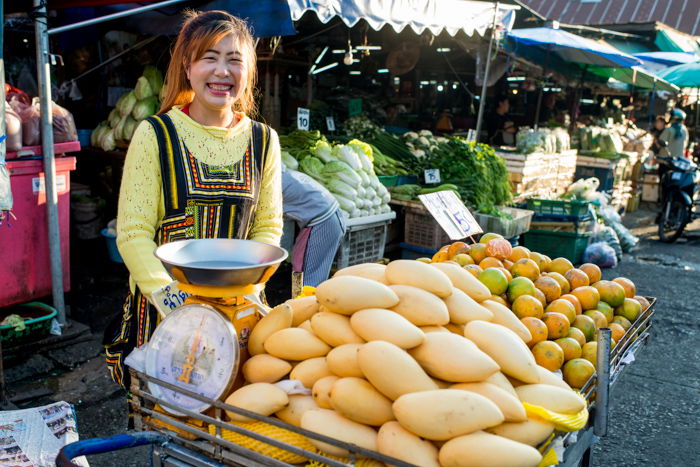 An environmental portrait of a mango seller standing at her market stall in Thailand