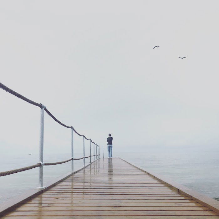 A person standing on a pier in the fog with birds flying overhead.