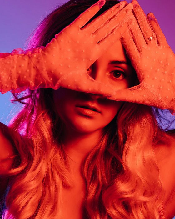 Editorial portrait of a woman with one eye peeking through a triangle made with her hands and colored lighting