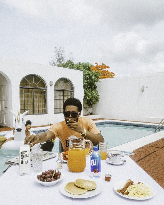 Editorial lifestyle image of a man eating breakfast at a table outside with a woman in a swimming pool behind him