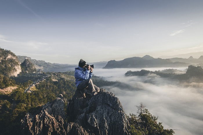 photographer sitting on a cliff with a mountain landscape in the background