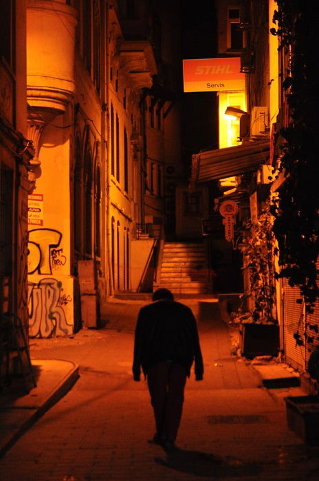 A raw style street photograph of a dark silhouette in an empty alleyway with their down with red and orange lighting