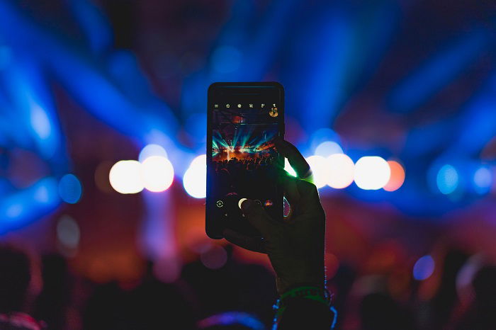 A hand holding up a smartphone with a camera app showing an event with a light display against a blurred background