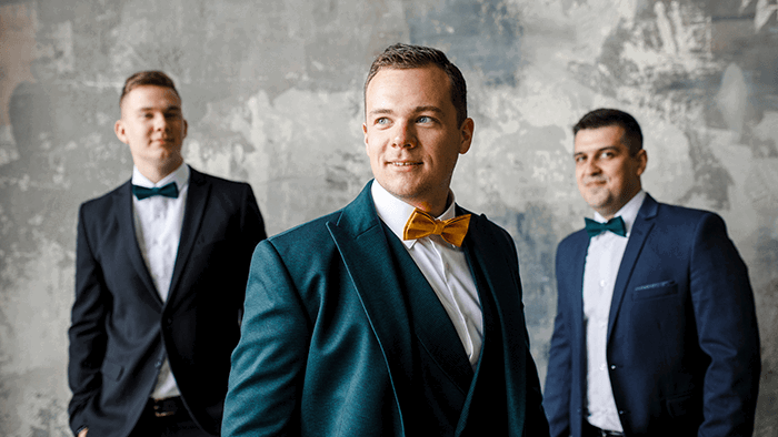 A groom wearing a bow-tie and suit with out-of-focus groomsmen standing behind him as an idea for photography posing