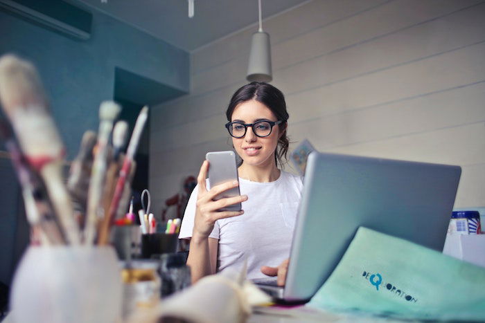 A woman looking at a smartphone while sitting behind a desk with a laptop and art supplies