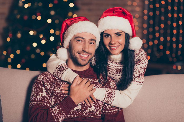 A couple wearing Christmas sweaters and Santa Clause hats posing for an indoor photoshoot