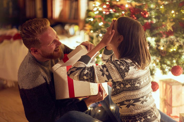 Christmas couple photoshoot ideas: a boyfriend opens the present box while the girlfriend covers her eyes with her hands 