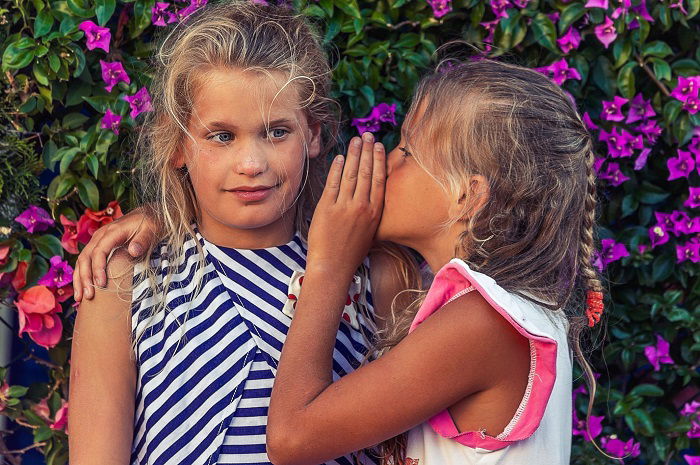 one girl whispering to another girl in front of flowers as sister photoshoot idea