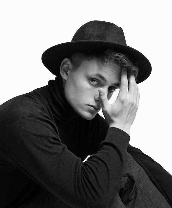 A black-and-white half-portrait a young male with a hat sitting and posing with his hand in front of his face and his fingers touching his hair