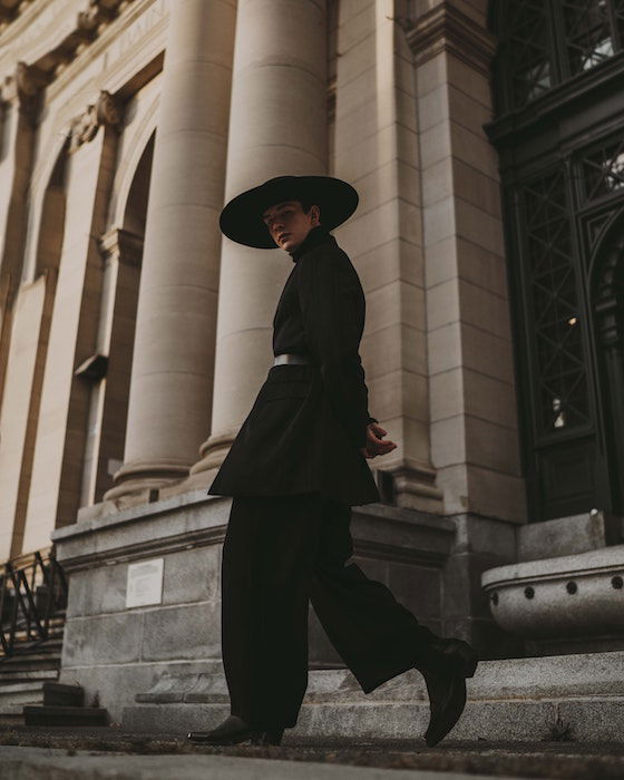 Man dressed in black walking in front of a building with hands behind his back as an example of model poses