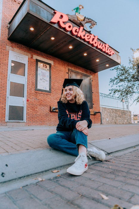 A graduate sitting on a crib in front of a restaurant with a grad cap on