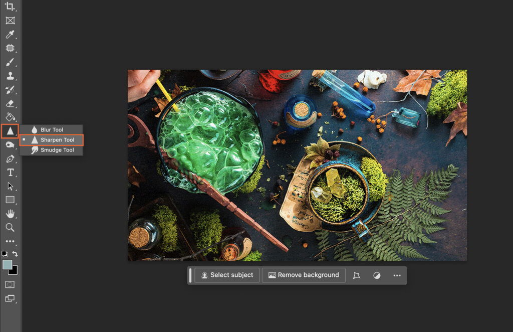 A photo editing software displaying a vibrant green beverage in a cup, surrounded by various other cups and bottles on a dark countertop, with a persons hand visible at the top left corner, holding a pen. 