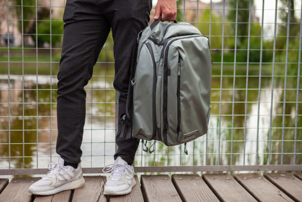 A person standing by a fence, holding an aqua-colored backpack and looking towards the camera. 